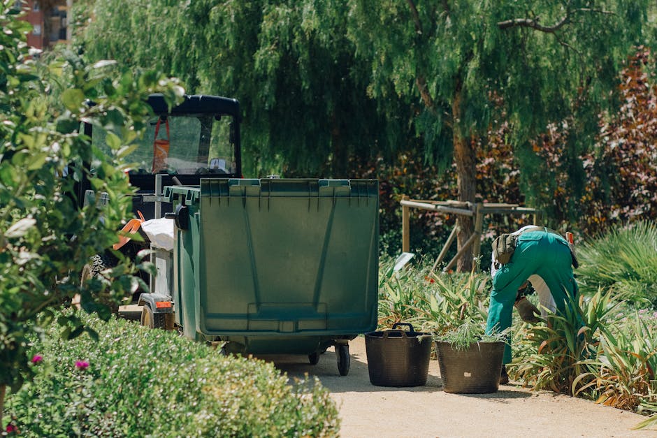 A gardener wearing teal coveralls is tending to a garden bed filled with tall, green, spiky plants, possibly ornamental or flowering varieties, on a sunny day. The gardener is bent over, actively prun