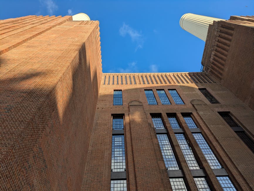 A low-angle view of a historic brick building in Battersea, with tall, narrow windows and architectural detailing, rising up towards a bright blue sky. Two large, cylindrical smokestacks are visible i