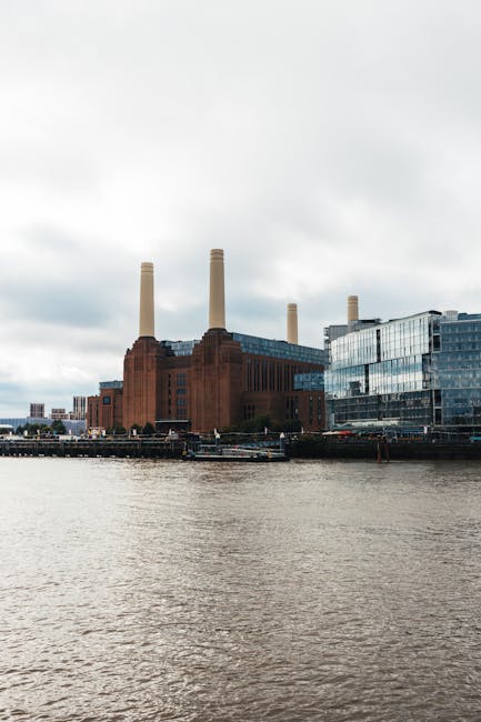 A low-angle view of a historic brick building in Battersea, with tall, narrow windows and architectural detailing, rising up towards a bright blue sky. Two large, cylindrical smokestacks are visible i
